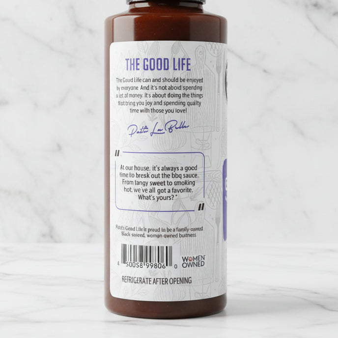 A bottle of 'The Good Life' barbecue sauce shown from the back, displaying the white label with blue text and a barcode, with other red-capped bottles blurred in the background.